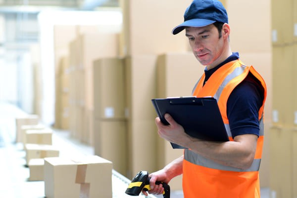 Warehouse worker in an orange safety vest scanning a box with an RF scanner while looking at a clipboard